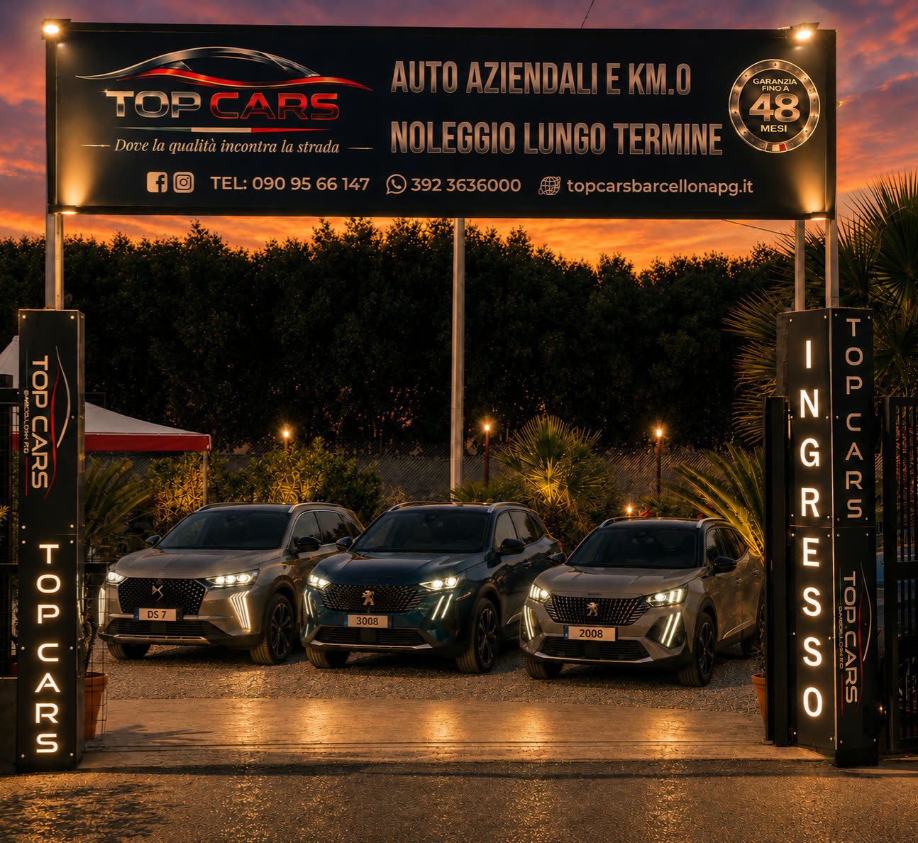 Il piazzale di Top Cars a Barcellona Pozzo di Gotto al tramonto: insegna illuminata e tre auto in esposizione